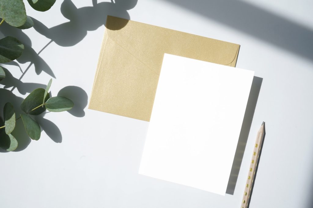 white printer paper and a brown envelope laying on a white background with a pencil and greenery. 100 envelope money saving challenge.