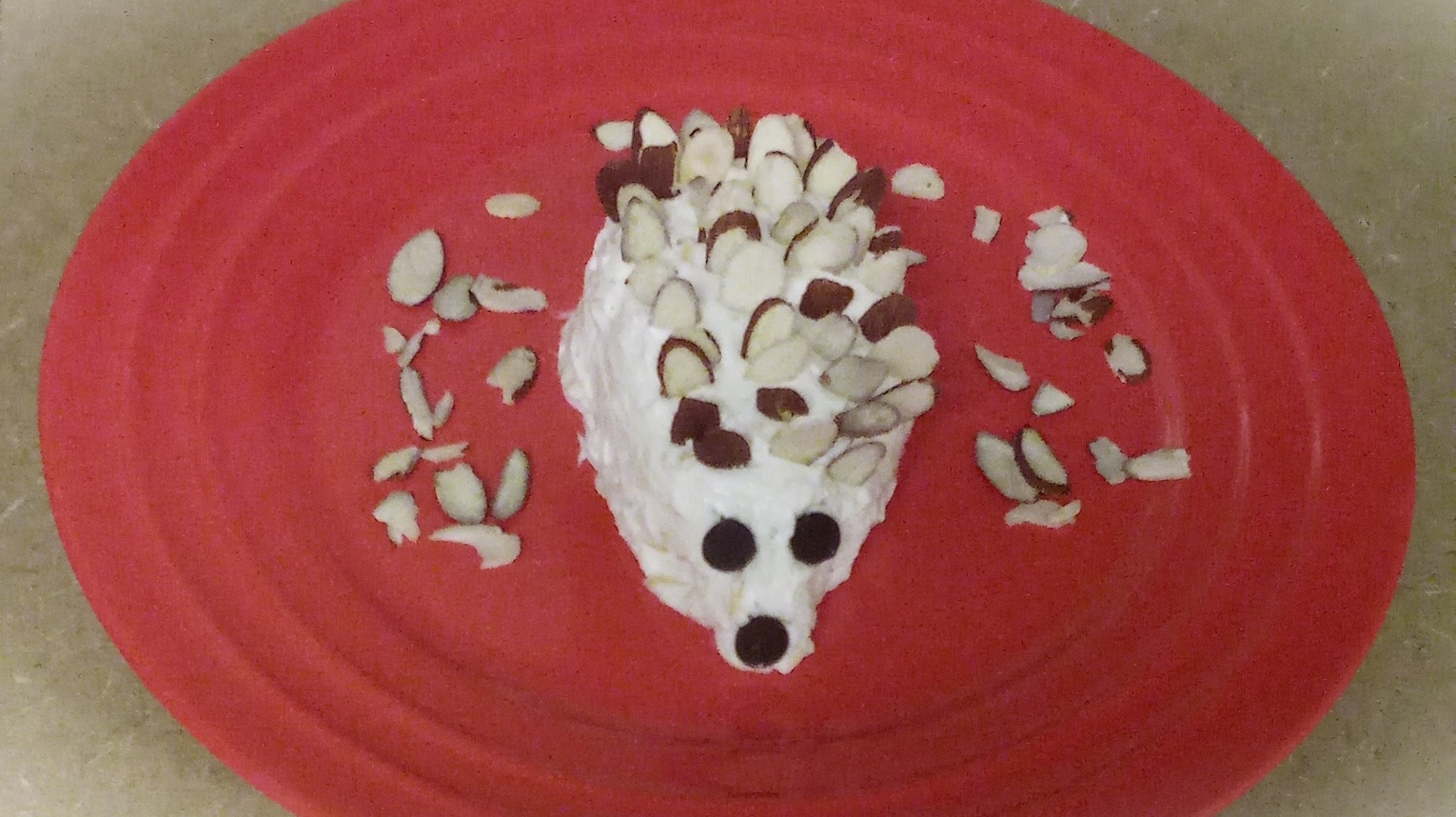 Hedgehog cheese ball on a red plate with slices of almonds laying around the cheese ball. Hedgehog Cheese ball