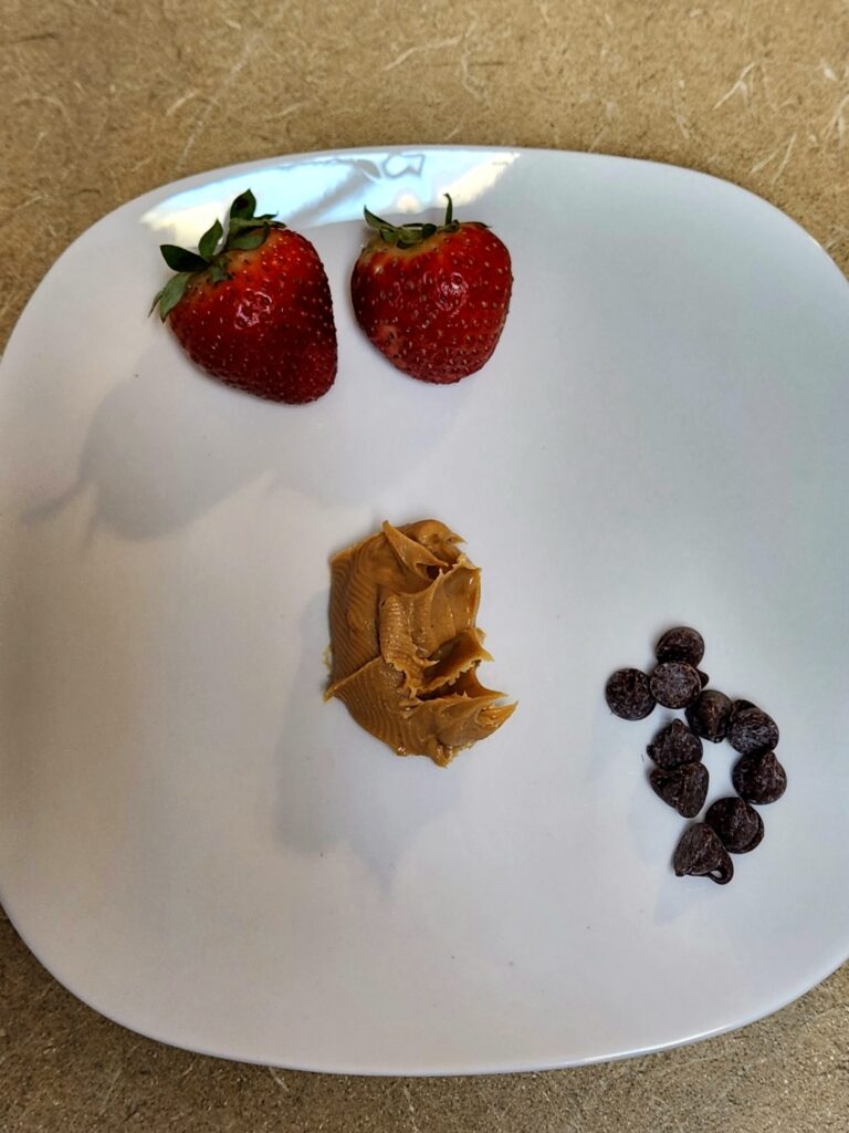 Waffle recipe, white plate with two strawberries, a scoop of peanut butter and some chocolate chips on it.